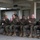 Commanding General's Quarterly Awards Ceremony A group of approximately nine military personnel in U.S. Marine Corps MARPAT camouflage uniforms are seated in a row on blue folding chairs, applauding during what appears to be a formal ceremony or event. They are positioned under a covered outdoor structure with concrete columns and beams. The group includes personnel of varying ranks, identifiable by their uniform insignia.