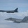 170214-N-XZ522-006 Two U.S. military aircraft are flying in formation over a cloud layer. The upper aircraft is a B-1B Lancer strategic bomber with tail code 'DY', and the lower aircraft is an F/A-18 Hornet or Super Hornet marked '303' with a Navy 'NE' tail code. This appears to be a joint USAF and U.S. Navy formation flight.