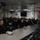 170214-N-YG414-167 A senior U.S. Navy officer in Navy Working Uniform (NWU) Type I digital blue camouflage is addressing a group of assembled sailors in what appears to be a briefing room or mess deck aboard a naval vessel. The audience consists of approximately 20-30 sailors seated at tables, attentively listening to the speaker. The setting suggests an official briefing, all-hands meeting, or command visit.