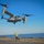 170214-N-NJ910-0256 A Bell Boeing V-22 Osprey tiltrotor aircraft is hovering low over a naval vessel's flight deck, marked with the number '00'. A flight deck crew member in a yellow jersey is directing the aircraft using hand signals. The scene takes place over open ocean under clear blue skies.
