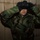 MCESG Gas Chamber A person wearing US military woodland camouflage BDU (Battle Dress Uniform) is in the process of donning or adjusting a black gas mask/protective mask. Both hands, covered in black gloves, are raised to hold the mask against their face/forehead. The subject is positioned against a concrete block wall in what appears to be an indoor or sheltered environment.