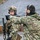 170213-N-N0901-002 Two individuals in MultiCam/OCP camouflage uniforms are engaged in what appears to be a training exercise at a shooting range or tactical training facility. One person, fully equipped with a helmet, ballistic eyewear, tactical vest, and rifle, is receiving directional instruction or guidance from a second person wearing hearing protection. The background shows a gravel berm typical of a military or law enforcement firing range.