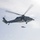 170214-N-WV703-163 A military helicopter is hovering over a ship's flight deck while conducting a hoist/rescue operation. A red litter (rescue stretcher) is suspended on a cable beneath the helicopter, with a deck crew member on the flight deck holding a guide line to control the load. The scene appears to be a medical evacuation or search-and-rescue training exercise at sea.