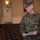 MCIOC Relief and Appointment A U.S. Marine in MARPAT digital camouflage uniform is speaking at what appears to be a formal military ceremony or briefing held in a banquet or conference hall. The Marine, identified by the name tape 'REAVES,' is wearing a medal consistent with the Legion of Merit and is holding papers, suggesting he is delivering remarks. An audience of seated military personnel is visible in the background.