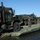 Bridge Company crosses the gap Military tactical trucks in olive drab camouflage are loaded onto a pontoon ferry or landing craft on a body of water. At least two uniformed personnel are visible managing the vehicles on the flat deck. The scene appears to depict a river-crossing or amphibious logistics operation.