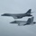 170214-N-XZ522-008 Two U.S. military aircraft are flying in formation against an overcast gray sky. The upper aircraft is a large swept-wing strategic bomber, and the lower is a twin-engine tactical fighter jet. Both aircraft appear to be in active flight during what appears to be a joint military operation or escort mission.