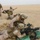 FASTCENT conduct live-fire training in CENTCOM AOR The image shows multiple military personnel in desert camouflage uniforms conducting what appears to be a live-fire training exercise in an arid desert environment. A prone shooter is operating a belt-fed machine gun positioned on green sandbags, with spent brass casings and linked ammunition visible nearby. Additional personnel are providing instruction or supervision, with one individual giving a hand signal in the background.