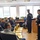 170209-N-VK873-004 A U.S. Navy officer and two enlisted sailors in dress uniforms are presenting or speaking to a group of young civilians, likely students, in what appears to be a school library or classroom setting. The officer, wearing a white-topped cover and gold-striped uniform, is gesturing while addressing the seated audience. This appears to be a community outreach or educational engagement event.