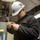 170214-N-DA693-008 A U.S. Navy sailor wearing a white hard hat marked '74' and safety glasses is working on a small electronic device or radio component. He is dressed in a black fleece jacket with a Navy eagle insignia and digital camouflage (NWU) trousers, indicating active duty military service. The setting appears to be an interior workspace aboard a naval vessel.