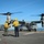 170214-N-NJ910-0221 A V-22 Osprey tiltrotor aircraft is being directed by flight deck crew aboard a naval vessel, likely an amphibious assault ship. Two personnel in yellow vests and helmets marked 'LSE' (Landing Signal Enlisted) are marshaling the aircraft on the flight deck. Additional helicopters and deck crew are visible in the background.