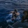 RIMPAC’s lost shots Military personnel in full combat gear are conducting a maritime water insertion or extraction operation in open ocean conditions at dusk or dawn. A central figure in desert camouflage uniform with a tactical helmet equipped with a green chemical light marker is reaching toward a vessel's boarding ladder while partially submerged. At least two additional armed personnel are visible — one in the water to the left and one on the vessel to the right.