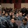 170215-N-JD834-023 A U.S. Navy briefing or all-hands call is taking place in an auditorium, with a speaker addressing a large audience of sailors in Navy Working Uniform (NWU) digital camouflage. One individual in civilian black clothing is standing, possibly asking a question or being recognized. The setting appears to be a military installation theater or lecture hall.
