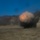 KMEP 17-8 The image captures an explosion occurring in an arid, open terrain surrounded by rocky hillsides. A large fireball with orange and gray tones is visible at ground level, accompanied by a significant black smoke cloud expanding outward. The surrounding landscape appears to be a dry, barren valley or quarry-like area.