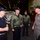 Rear Adm. Kreitz meets with USS Nimitz triad Four U.S. Navy personnel are engaged in conversation inside what appears to be a ship's hangar bay, likely aboard an aircraft carrier. Three individuals wear navy blue or olive green flight jackets with caps bearing what appears to be 'Nimitz' insignia, while the fourth wears a gray coverall uniform with a star insignia indicating flag officer rank. The setting features covered equipment or aircraft in the background with industrial lighting overhead.