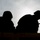 KMEP 17-8 A backlit silhouette photograph shows two individuals working on what appears to be a construction project. The figures are rendered entirely in dark silhouette against an overcast, bright sky. One person is actively using a hammer while the other appears to be observing or assisting.