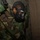 MCESG Gas Chamber A military service member wearing woodland camouflage BDUs and a black M40-series gas mask is positioned against a concrete block wall. The individual appears to be in a training exercise, likely a CBRN (Chemical, Biological, Radiological, Nuclear) drill or gas chamber exercise. Black tactical gloves are also visible as part of the protective equipment ensemble.