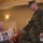 MCIOC Relief and Appointment A U.S. Marine in digital camouflage uniform is leaning down to shake hands with an elderly seated woman who is holding a wrapped bouquet of flowers. The setting appears to be a formal indoor ceremony or event in a well-appointed room. The Marine is wearing a medal on his chest, suggesting this is an official military ceremony.