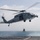 170214-N-WV703-103 A U.S. Navy helicopter, marked with the number '45' and labeled 'NAVY', hovers over the deck of a naval vessel at sea while conducting a hoist/rescue operation. A person in flight gear is suspended below the aircraft on a hoist cable. The scene appears to be a training or operational exercise conducted over open ocean.