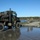 Bridge Company crosses the gap Military personnel are conducting a river crossing operation using a pontoon ferry or floating bridge. A large military tactical truck (appears to be an LVSR or similar USMC heavy logistics vehicle) is being guided onto or off of the floating bridge structure over a body of water. Several uniformed soldiers/Marines are directing the vehicle and managing the crossing operation.
