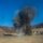 KMEP 17-8 The image shows a large explosion or controlled detonation occurring in an open, arid valley surrounded by hills with sparse, dry vegetation. A tall column of dark black smoke and debris is rising from ground level against a clear blue sky. The scene appears to be a demolition, mining blast, or ordnance disposal event in a remote outdoor location.