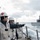 170212-N-HI376-011 Naval personnel in military uniforms and safety gear stand on the deck of a warship observing another vessel nearby. One sailor wearing a white helmet with a red cross (medical insignia) and another pointing toward the adjacent ship suggest a replenishment-at-sea or coordination operation. The scene takes place in rough open-ocean conditions under overcast skies.