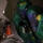 Chemical, Biological, Radiological and Nuclear Marines Participate Advanced Training A person wearing a full green hazmat/CBRN protective suit with a large face shield visor is leaning over a laboratory bench. The suit has visible yellow labels and markings including what appears to 'CHEM' text. The scene appears to be a hazardous materials investigation or training exercise in a dimly lit room.
