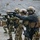 170213-N-N0901-005 A group of heavily equipped tactical operators are conducting pistol marksmanship training at an outdoor shooting range. Multiple personnel are lined up in a firing line, aiming handguns downrange in a coordinated drill. The setting appears to be a gravel-surfaced range with a berm backdrop, consistent with a controlled military or law enforcement training environment.