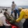 170214-N-WV703-133 A medical evacuation drill or casualty response exercise is taking place on the flight deck of a naval vessel at sea. Multiple personnel in different colored vests and helmets with hearing protection are handling a simulated casualty on a stretcher/litter. One person wearing a white vest with a Red Cross symbol is among the responders, indicating medical personnel involvement.