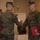 MCIOC Relief and Appointment A military awards ceremony is taking place in an indoor venue, likely a banquet hall or conference room. Two U.S. Marines in MARPAT digital camouflage uniforms are shaking hands, with one Marine wearing a medal on his chest and holding a red award folder. A female Marine is visible in the background also holding a red folder.