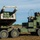 10th Marine Regiment HIMARS Firing Excercise The image shows a military multiple rocket launch system (MLRS) vehicle, specifically appearing to be an M270 or HIMARS-type system mounted on a heavy tactical truck chassis, positioned in an open field. The launcher pod is elevated and angled, suggesting a pre-fire or post-fire position. At least one uniformed military personnel is visible in the vehicle's cab area.