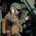 10th Marine Regiment HIMARS Firing Excercise A military service member in MARPAT digital camouflage uniform is positioned at the entrance of a military vehicle, likely a JLTV or HMMWV. The soldier is wearing a combat helmet with a communications headset and holding a handset to their ear while gripping the steering wheel. They are equipped with a tactical backpack and appear to be communicating via radio.