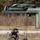 A U.S. Marine in full combat gear and flight helmet is working with a rocket pod assembly, likely part of a HIMARS or similar multiple launch rocket system (MLRS) reload operation. The equipment is labeled 'INERT TRAINER' and 'DO NOT PUSH,' indicating this is a training exercise rather than a live-fire event. Rigging lines from an overhead crane or lift mechanism are being managed by the Marine to position the rocket pod.