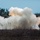 10th Marine Regiment HIMARS Firing Excercise The image captures a large explosion or detonation occurring in an open field near a treeline. A massive white and light-orange smoke cloud billows upward, with what appears to be a small dark projectile or debris fragment visible mid-air within the smoke. The setting appears to be a rural or semi-arid landscape under clear blue skies.