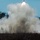 10th Marine Regiment HIMARS Firing Excercise The image captures a missile or rocket at the moment of launch, rising above a large cloud of white exhaust smoke and propellant gases. The launch appears to be occurring in a rural or semi-wooded area, with low vegetation and trees visible in the background. The projectile is clearly visible at the top of the smoke plume with a bright ignition flame.