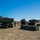 10th Marine Regiment HIMARS Firing Excercise The image shows multiple M270 Multiple Launch Rocket System (MLRS) or M142 HIMARS-type vehicles deployed in an open field during what appears to be a military training exercise. Several soldiers in camouflage uniforms are working around the vehicles, with rocket pod containers visible on the ground between the launchers. The setting appears to be a flat, dry training range with sparse vegetation and a clear blue sky.