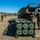 The image shows U.S. Marines conducting a rocket artillery exercise in an open field, likely a training environment. A Multiple Launch Rocket System (MLRS) or similar vehicle is visible alongside a reload pod container marked '789' holding six rocket tubes. Personnel in MARPAT camouflage uniforms and combat gear are actively working around the equipment.