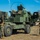 Military personnel in MARPAT camouflage uniforms are conducting operations around what appears to be an M142 HIMARS (High Mobility Artillery Rocket System) or similar multiple launch rocket system on an open field. At least three service members are visible, with one in a turret position, one working near the launcher pod, and one holding what appears to be a remote firing cable or communication line. The setting appears to be a training exercise at a military installation.