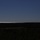 10th Marine Regiment HIMARS Firing Excercise A rocket or missile is captured mid-launch, ascending diagonally across a clear deep blue sky, leaving a distinct white exhaust contrail behind it. The launch appears to originate from the lower right of the frame, near a coastal area. The foreground is heavily shadowed, showing a dark landscape of low vegetation and what appears to be a road or path.