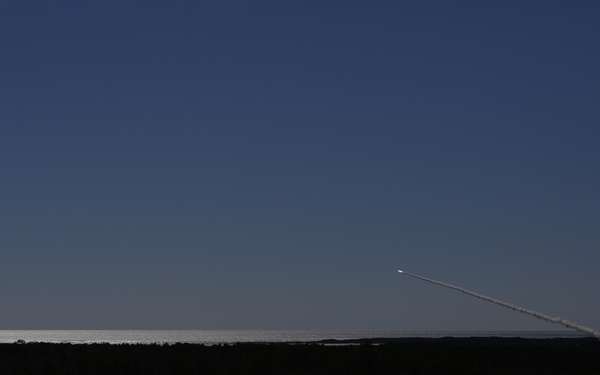 A rocket or missile is captured mid-launch, ascending diagonally across a clear deep blue sky, leaving a distinct white exhaust contrail behind it. The launch appears to originate from the lower right of the frame, near a coastal area. The foreground is heavily shadowed, showing a dark landscape of low vegetation and what appears to be a road or path.