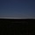 10th Marine Regiment HIMARS Firing Excercise A nighttime or twilight photograph capturing a rocket or missile launch trail arcing across a dark blue sky. The bright exhaust plume originates from the right side of the frame near the horizon and extends upward and to the left in a curved trajectory. The foreground is dominated by a dark, silhouetted landscape of trees and flat terrain.
