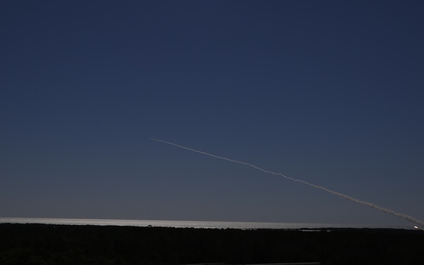 A nighttime or twilight photograph capturing a rocket or missile launch trail arcing across a dark blue sky. The bright exhaust plume originates from the right side of the frame near the horizon and extends upward and to the left in a curved trajectory. The foreground is dominated by a dark, silhouetted landscape of trees and flat terrain.
