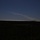 10th Marine Regiment HIMARS Firing Excercise The image shows a rocket or missile launch captured from a distance at dusk or twilight, with a prominent exhaust/vapor trail arcing across the sky from right to left. The foreground is dominated by a dark, silhouetted treeline and flat terrain. A bright point of light, likely the rocket itself, is visible near the right side of the trail.