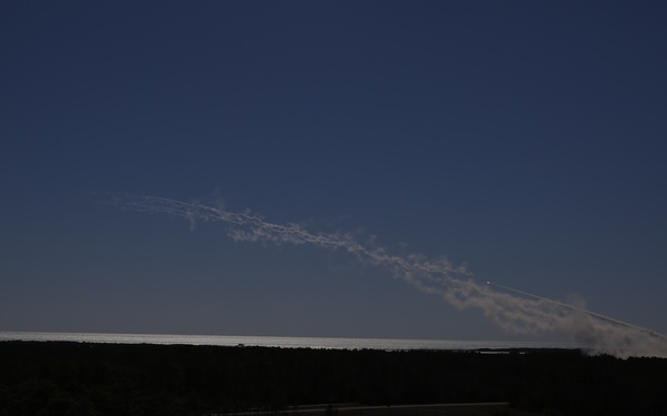 The image shows a rocket or missile launch captured from a distance at dusk or twilight, with a prominent exhaust/vapor trail arcing across the sky from right to left. The foreground is dominated by a dark, silhouetted treeline and flat terrain. A bright point of light, likely the rocket itself, is visible near the right side of the trail.