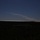 10th Marine Regiment HIMARS Firing Excercise A dark landscape photograph showing a wide, flat terrain covered with dense vegetation in the foreground, silhouetted against a deep blue sky. A prominent diagonal white plume or trail extends from the right side of the horizon upward toward the upper left of the frame. The source of the plume appears to originate near the horizon line on the right side, possibly from a fire, industrial source, or rocket launch.