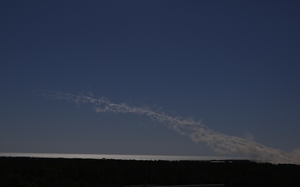 A dark landscape photograph showing a wide, flat terrain covered with dense vegetation in the foreground, silhouetted against a deep blue sky. A prominent diagonal white plume or trail extends from the right side of the horizon upward toward the upper left of the frame. The source of the plume appears to originate near the horizon line on the right side, possibly from a fire, industrial source, or rocket launch.