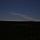 A dark landscape photograph showing a wide, flat terrain covered in dense vegetation or forest in the foreground. A prominent white smoke or exhaust plume extends diagonally across the upper portion of the image against a deep blue sky, originating from the right side of the horizon. The source of the plume appears to be on or near the ground at the right edge of the frame.