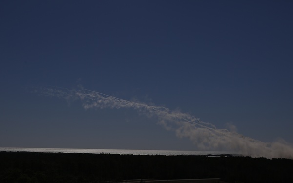 A dark landscape photograph showing a wide, flat terrain covered in dense vegetation or forest in the foreground. A prominent white smoke or exhaust plume extends diagonally across the upper portion of the image against a deep blue sky, originating from the right side of the horizon. The source of the plume appears to be on or near the ground at the right edge of the frame.
