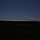 The image shows multiple white smoke trails arcing across a dark blue twilight sky, originating from the right side of the frame near the horizon. The foreground is heavily shadowed, revealing a dark treeline and flat terrain. The trails suggest multiple projectiles or rockets launched in rapid succession from a single origin point.