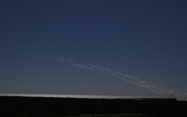 The image shows multiple white smoke trails arcing across a dark blue twilight sky, originating from the right side of the frame near the horizon. The foreground is heavily shadowed, revealing a dark treeline and flat terrain. The trails suggest multiple projectiles or rockets launched in rapid succession from a single origin point.