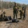 10th Marine Regiment HIMARS Firing Excercise Three military personnel in MARPAT camouflage uniforms and helmets are operating camera and video equipment in an open field during what appears to be a documentation or media coverage mission. One soldier stands holding a DSLR camera with a telephoto lens, another kneels sorting through equipment bags, and a third operates a video camera mounted on a tripod. The setting appears to be a military training area with a tree line in the background.